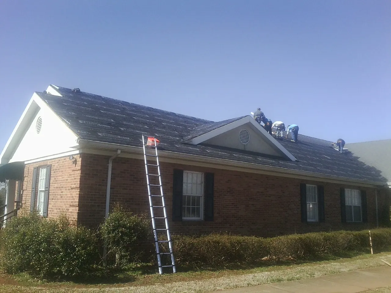 Crew tearing shingles off a brick ranch roof with a ladder set — Metal Roof Repair work by our crew in Candor